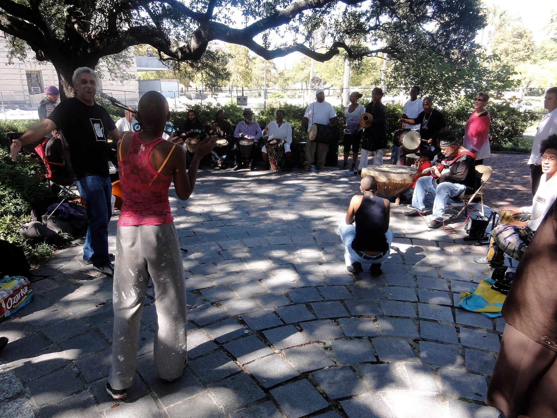 Un gruppo di persone si riunisce sotto una grande quercia a Congo Square, suonando tamburi, ballando e facendo musica in una giornata di sole.