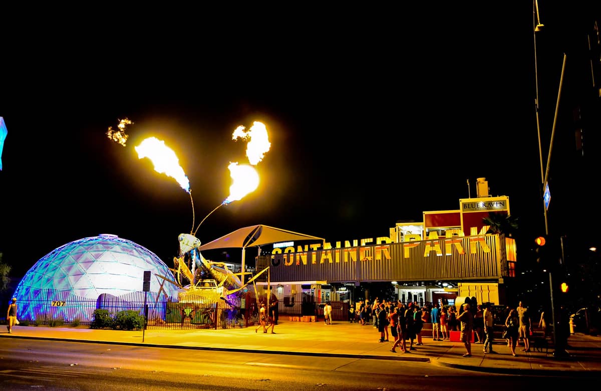 Vista notturna dell'ingresso del Downtown Container Park a Las Vegas, con la scultura di una mantide religiosa in fiamme, la cupola illuminata e la folla radunata fuori.