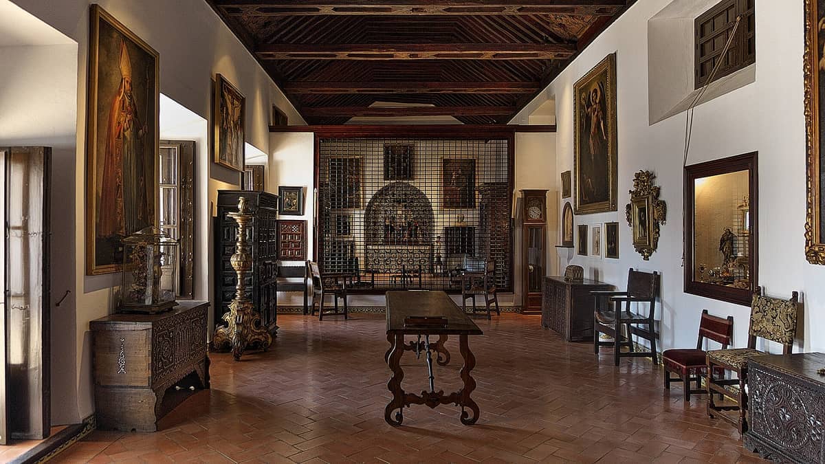 ภายในของ Convento de Santa Paula ที่มีภาพวาดทางศาสนา เฟอร์นิเจอร์ไม้โบราณ และแสงธรรมชาติอ่อนๆ ส่องสว่างห้องประวัติศาสตร์ที่ปูกระเบื้อง
