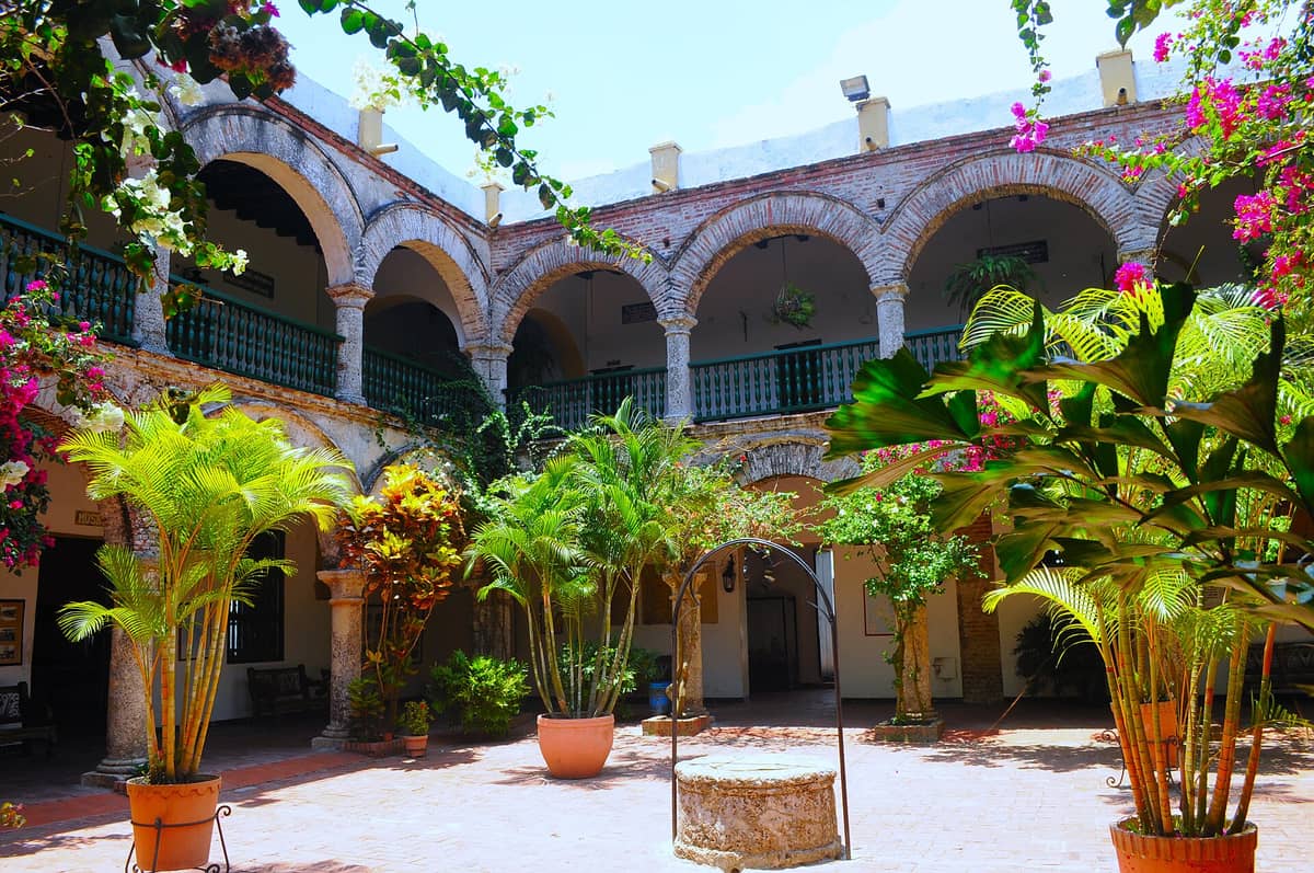 Bright inner courtyard of Cartagena's Convento de Santa Cruz de la Popa, showing colonial arches, potted plants, and a central stone well under blue sky.
