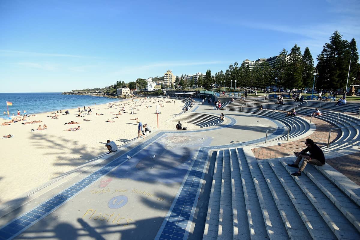 Wide-angle view of Coogee Beach showing sandy shores, blue ocean, curved concrete steps, sunbathers, and trees alongside low-rise buildings under clear skies.