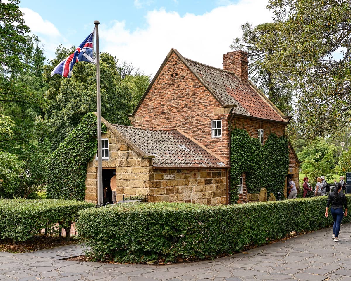 Cooks' Cottage, una pequeña casa de ladrillo y piedra del siglo XVIII cubierta de hiedra, con una bandera de la Unión Jack, rodeada de árboles y jardines en los Fitzroy Gardens de Melbourne.