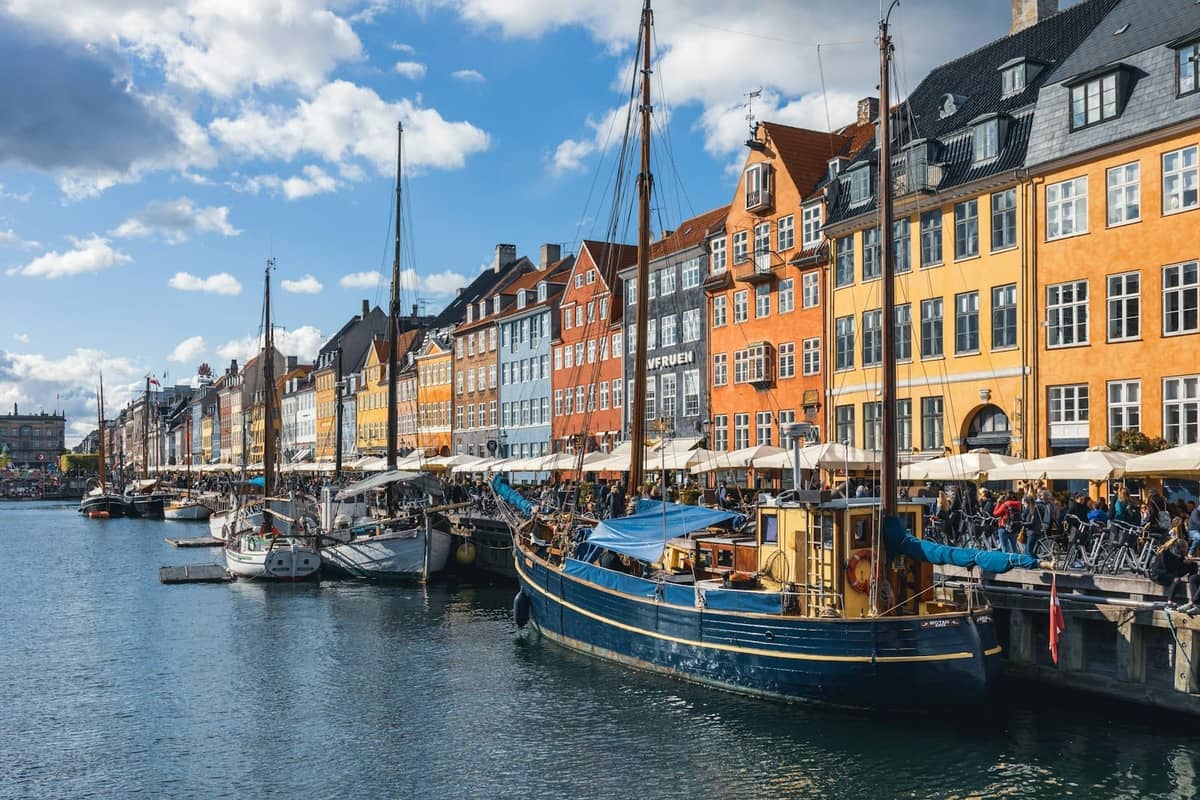 Kolorowe zabytkowe budynki i łodzie wzdłuż kanału Nyhavn w Kopenhadze, z ludźmi spacerującymi i jedzącymi na zewnątrz w słoneczny dzień.