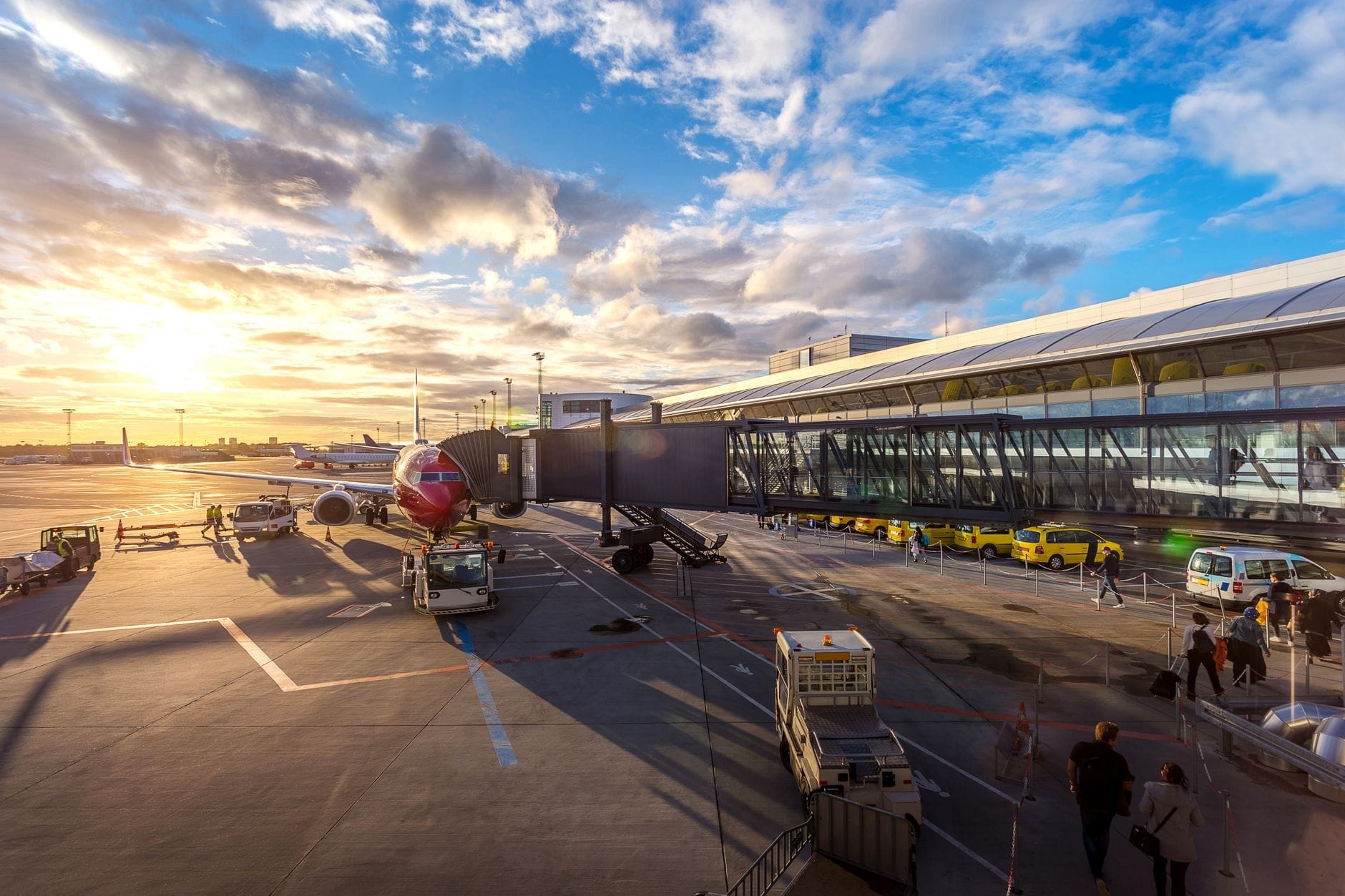 Aeroporto de Copenhague ao amanhecer com um avião no portão, táxis amarelos, terminal e passageiros caminhando no asfalto sob um céu vibrante.