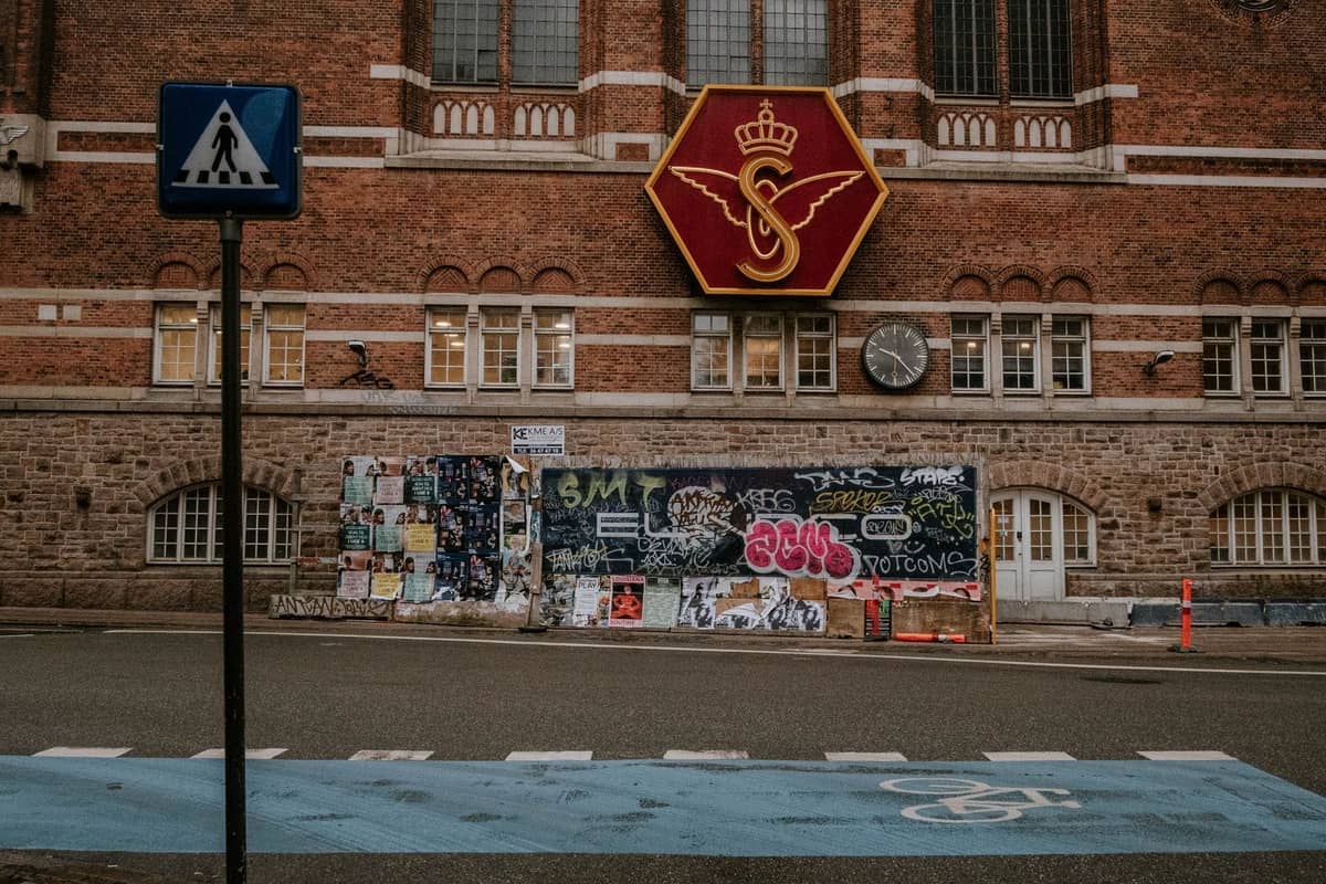 Widok z poziomu ulicy na historyczny ceglany budynek stacji w Kopenhadze z dużym szyldem skrzydlatego koła DSB i zegarem na fasadzie.