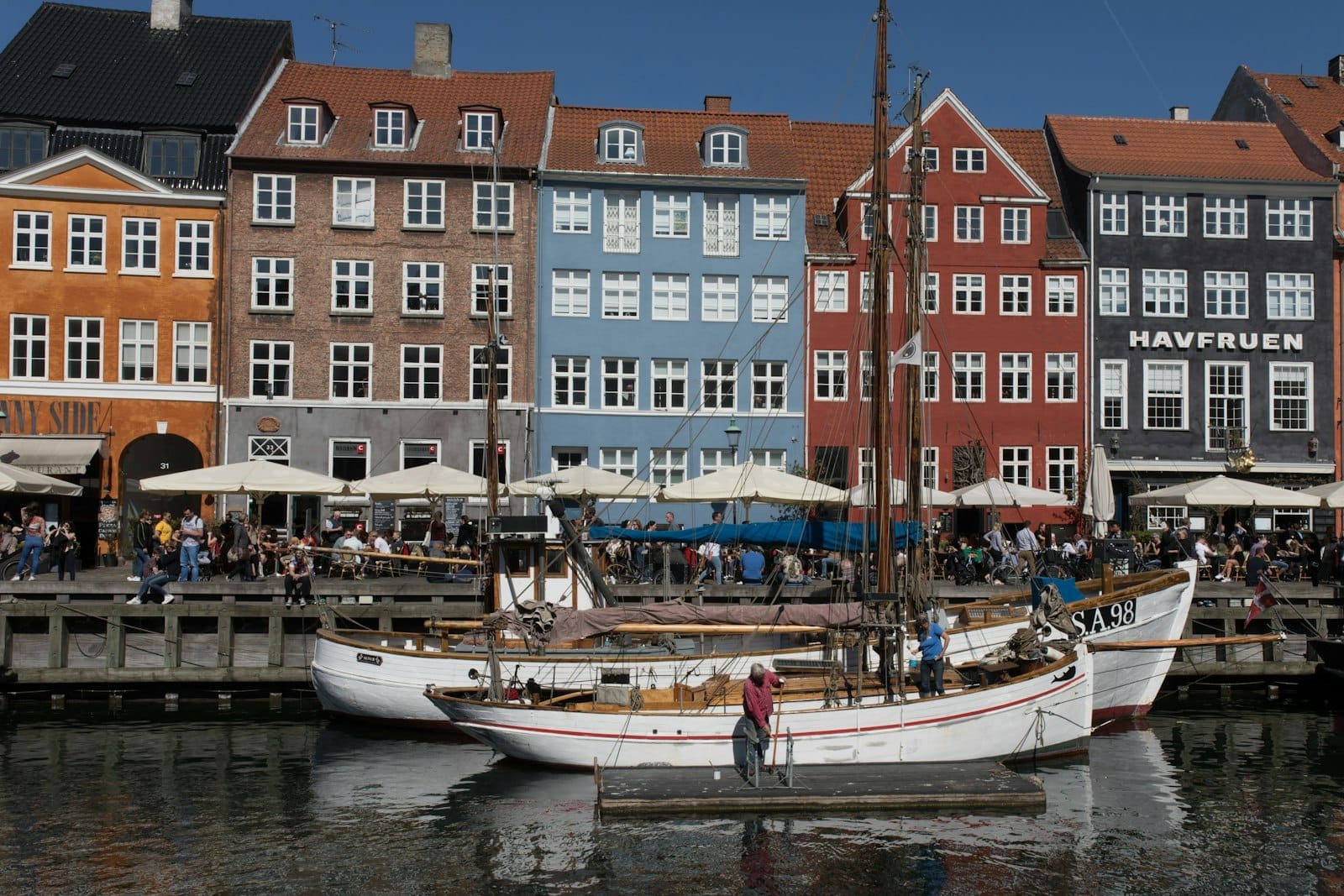 Prédios históricos coloridos e barcos ao longo do canal Nyhavn em Copenhague num dia ensolarado, com muita gente aproveitando cafés e passeios turísticos.