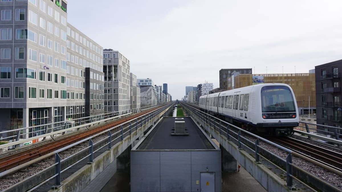Train de métro de Copenhagen circulant sur des voies surélevées avec des bâtiments urbains modernes de chaque côté, sous un ciel nuageux.