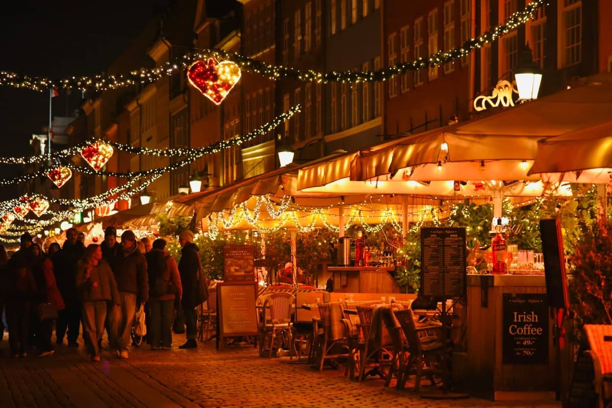 Kopenhagener Straße mit Weihnachtsbeleuchtung, Restaurantterrasse unter festlichen Dekorationen, Menschen beim Spazieren und Speisen, gemütliche Feiertagsatmosphäre.