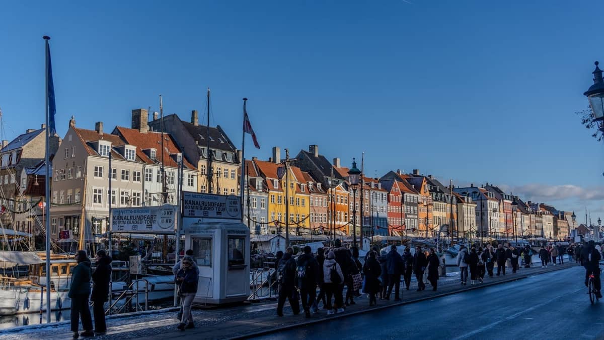 Menschenmassen schlendern an Kopenhagens bunter Nyhavn-Uferpromenade an einem frischen Wintertag entlang, Reihen historischer Gebäude unter klarem blauem Himmel.