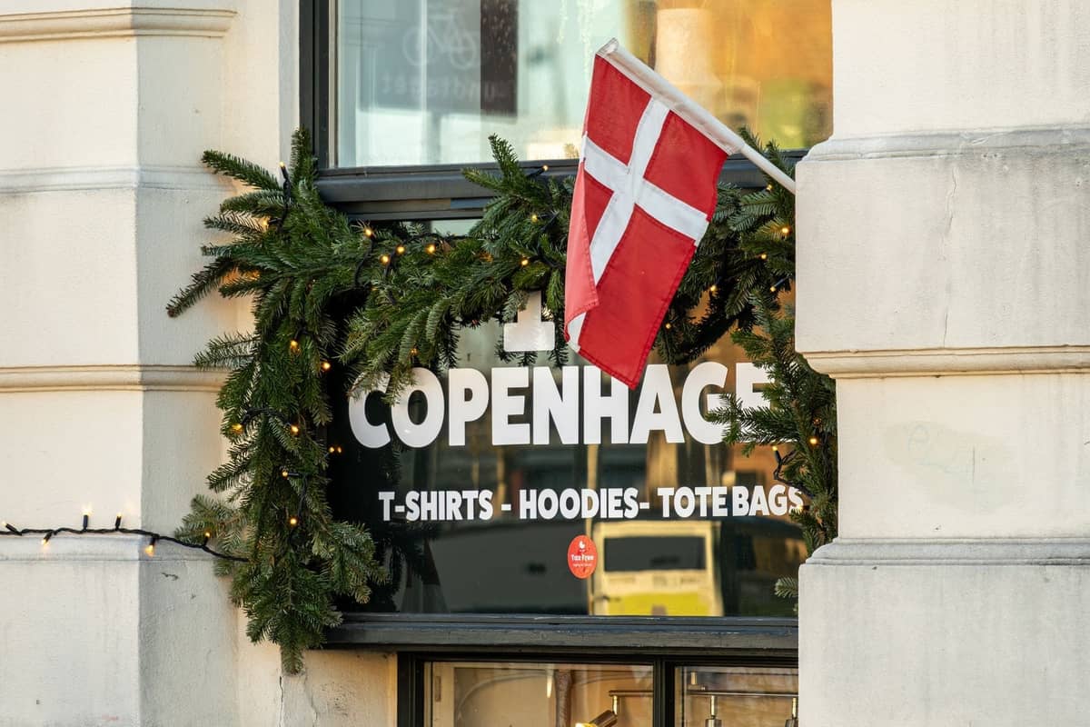 Außenansicht eines Souvenirladen in Kopenhagen, dekoriert mit einer dänischen Flagge und festlicher Girlande, der T-Shirts, Hoodies und Tragetaschen verkauft.