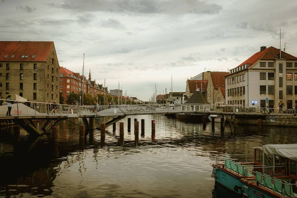 Scène de canal à Copenhague avec un pont piétonnier moderne, des bâtiments historiques aux toits rouges, des bateaux et un paysage urbain sous un ciel nuageux.