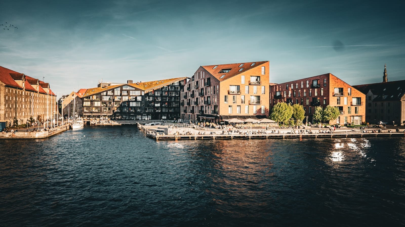 Edifícios modernos à beira-mar em Copenhague durante a hora dourada, exibindo a arquitetura dinamarquesa característica com reflexos na água e uma atmosfera vibrante e convidativa.