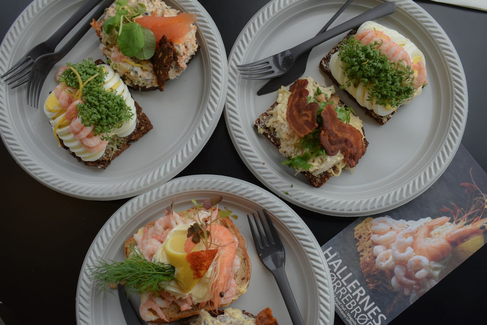 Quatro pratos de smørrebrød dinamarquês tradicional com diferentes coberturas, vistos de cima sobre uma mesa, mostrando a colorida e apetitosa culinária de Copenhague.
