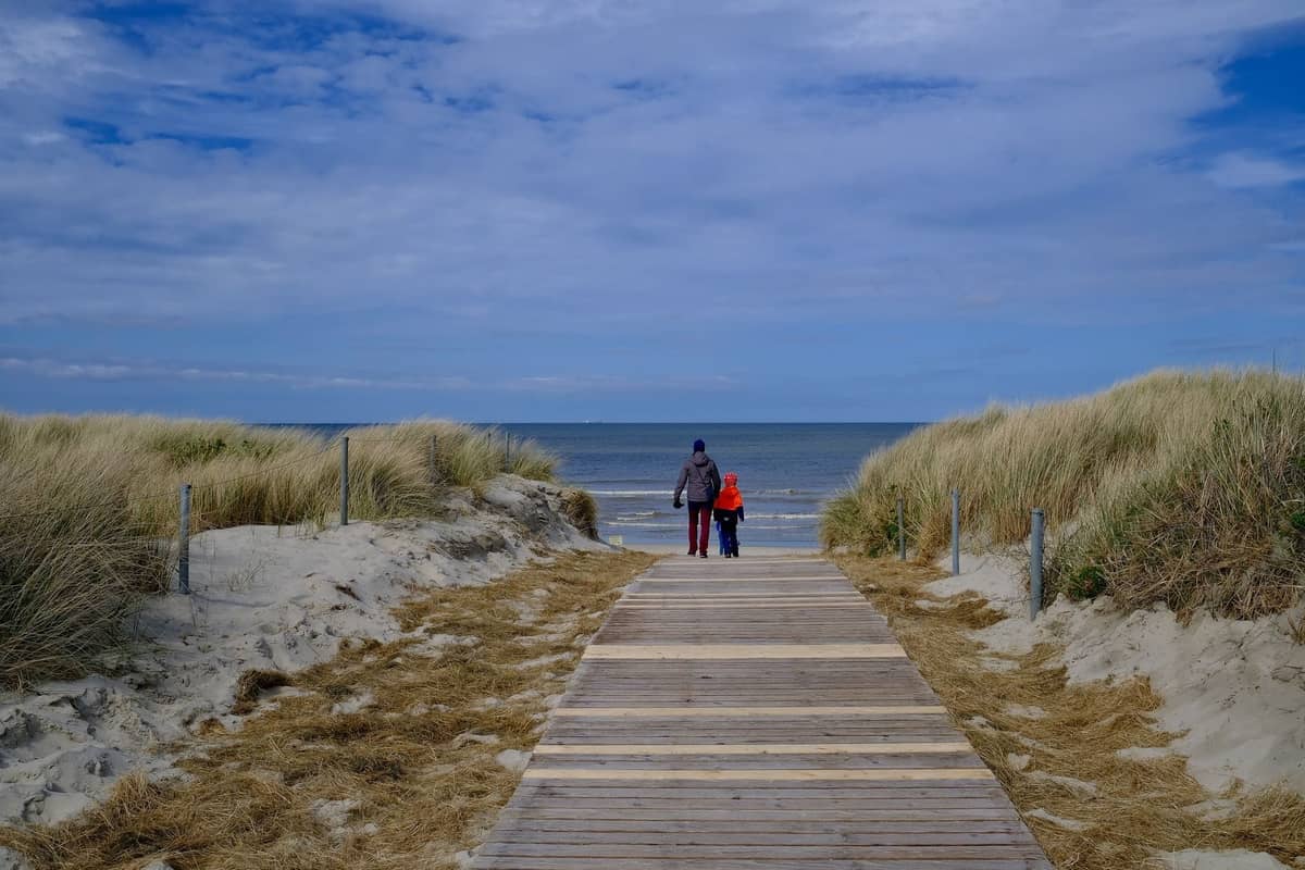 Due persone con giacche camminano verso una spiaggia deserta su una passerella di legno, con cielo nuvoloso e riva fredda e vuota.