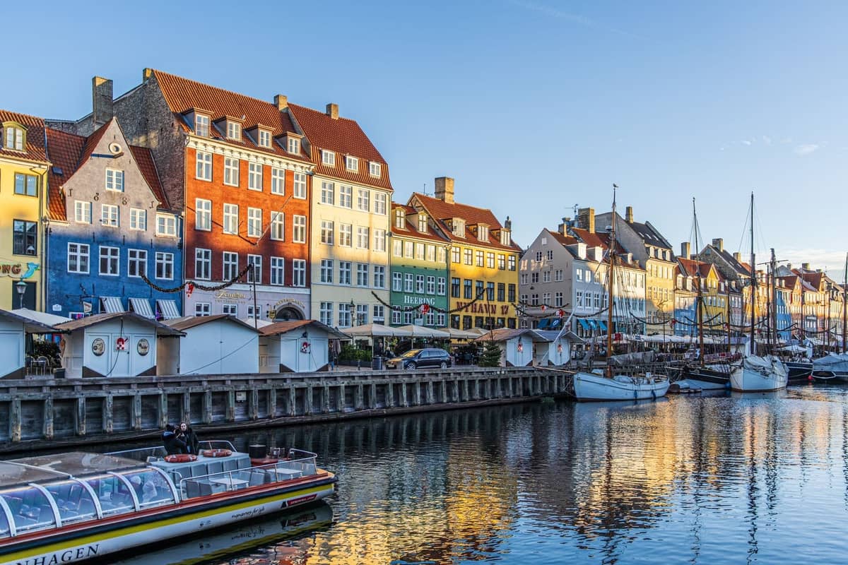La luminosa luce solare estiva illumina le colorate case storiche e le barche di Nyhavn a Copenaghen, con cieli azzurri limpidi e persone che godono del lungomare.