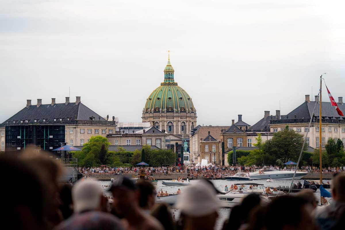 Grande folla estiva sul lungofiume di Copenaghen con barche e la cupola della Marble Church visibile sullo sfondo.