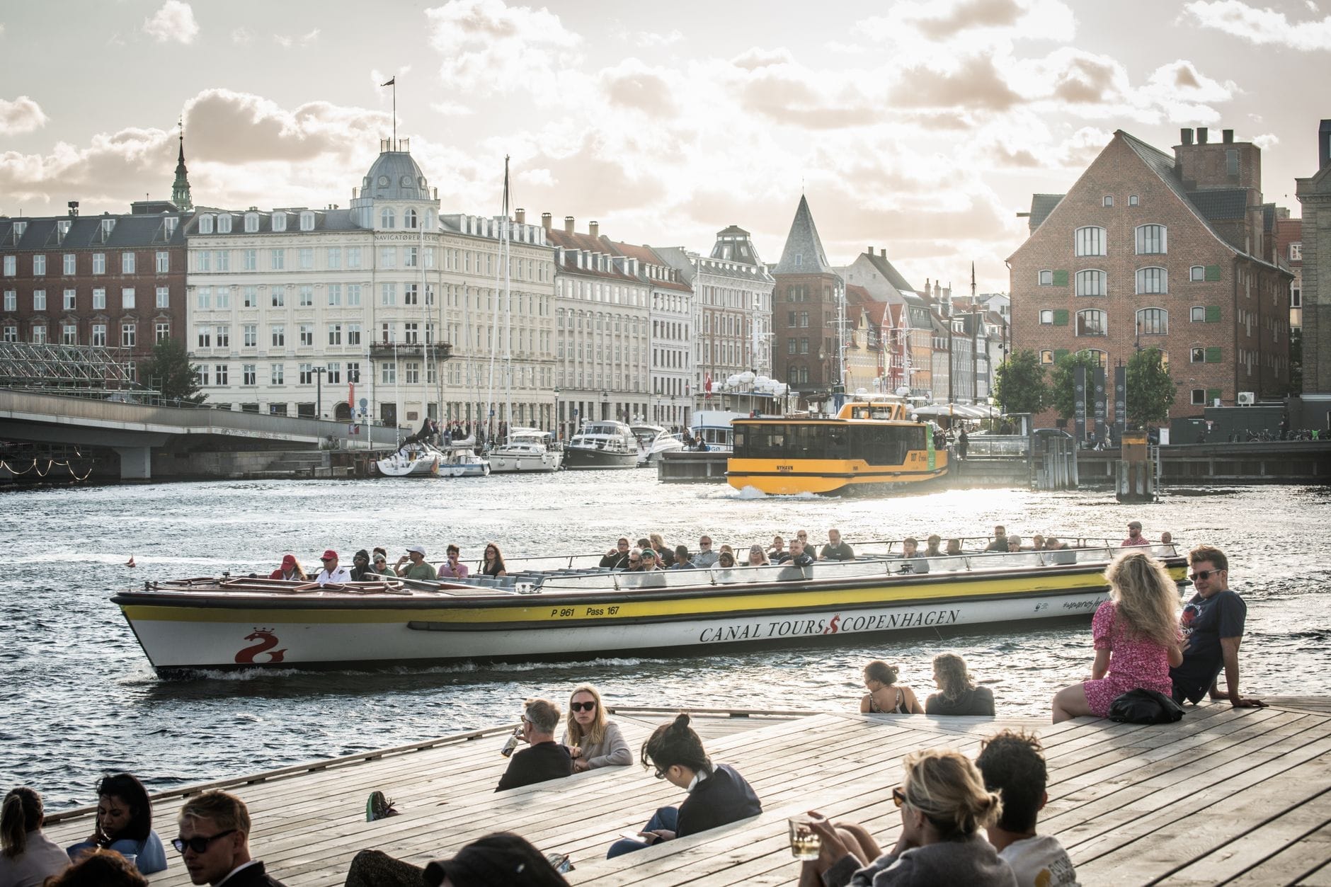 Pessoas relaxando em um deck de madeira em Copenhague observando barcos turísticos no canal, com prédios históricos ao fundo em um ensolarado dia de verão.