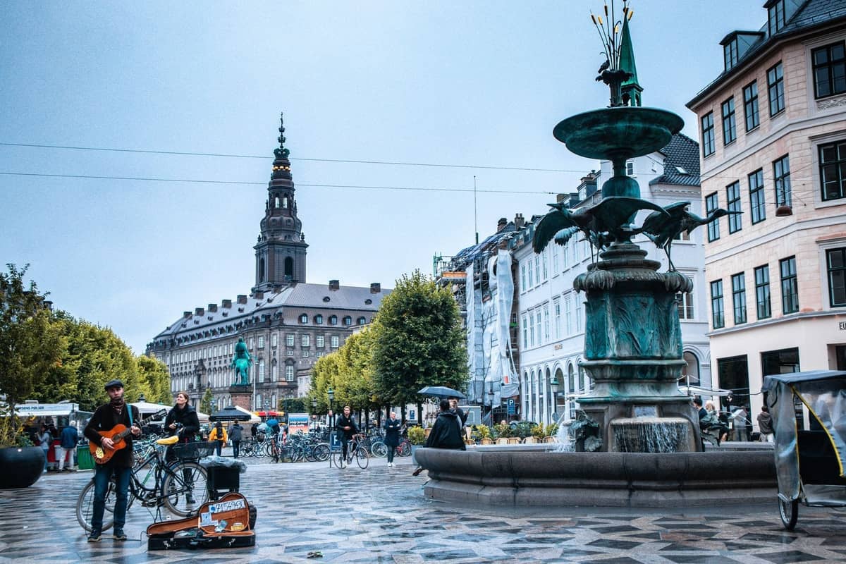 Stadtplatz in Kopenhagen mit einem Gitarre spielenden Musiker, Brunnen, historischen Gebäuden und Passanten an einem bewölkten Tag.
