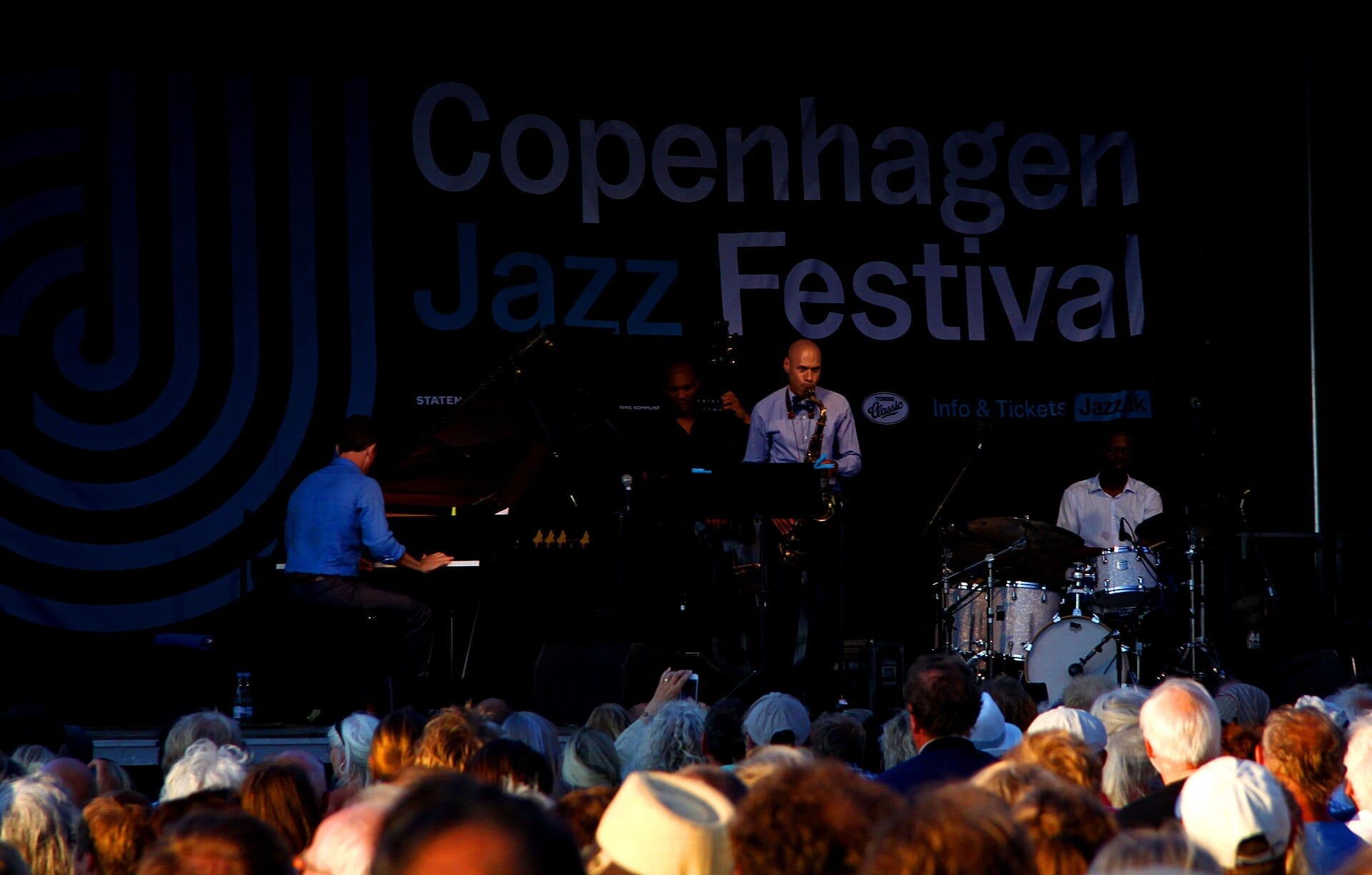 Banda de jazz se apresentando no palco do Copenhagen Jazz Festival, com o público assistindo sob as luzes da noite e o banner do evento ao fundo.