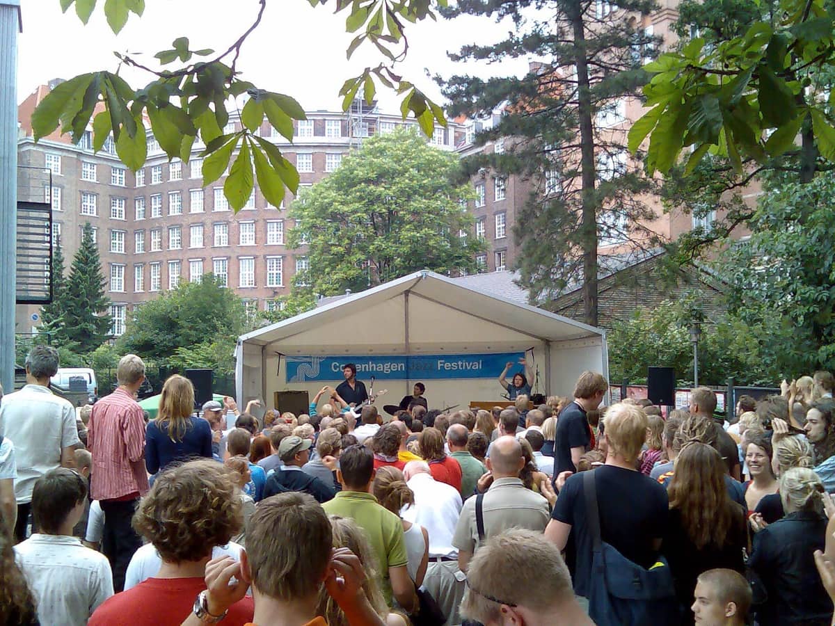 Eine große Menschenmenge versammelt sich vor einer Freilichtbühne beim Copenhagen Jazz Festival, umgeben von üppigen Bäumen und historischen Backsteingebäuden.