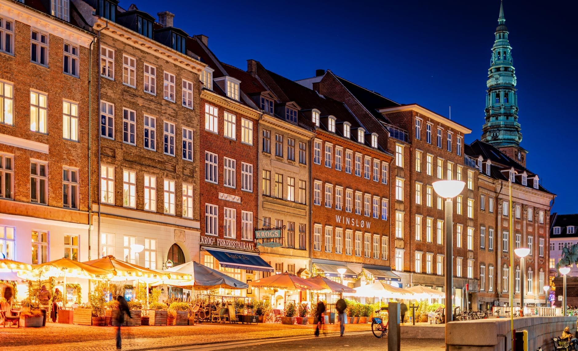 Vista noturna de uma rua movimentada de Copenhague repleta de bares e restaurantes animados, com prédios históricos coloridos e pessoas curtindo a vibrante vida noturna da cidade.