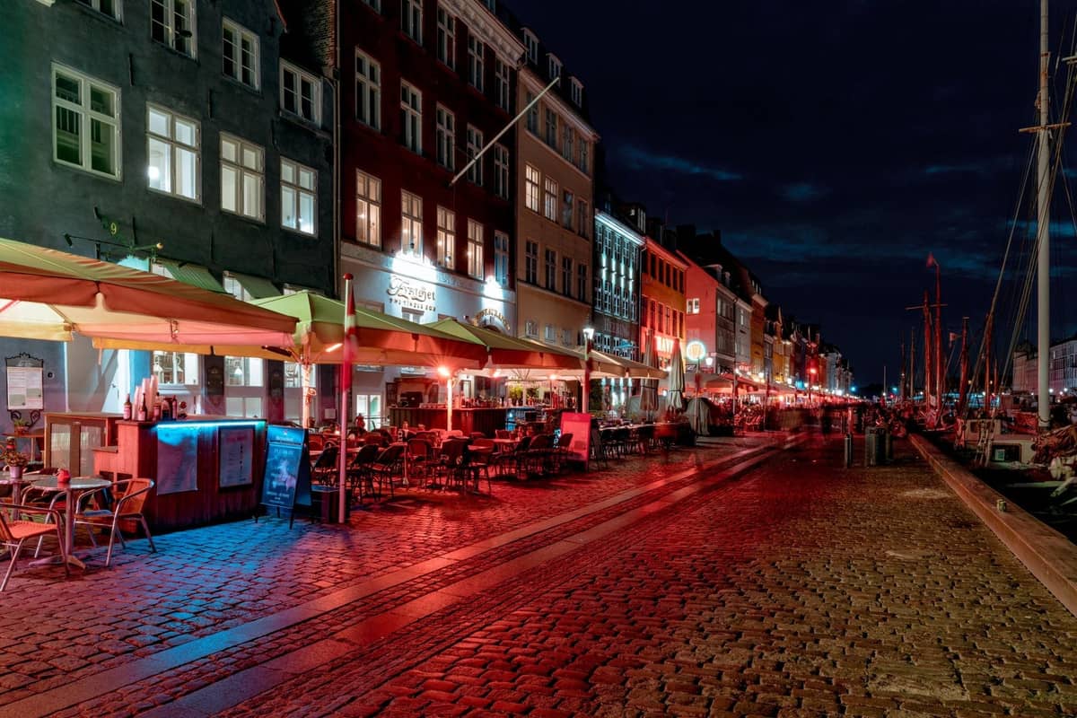 Kopenhag'daki Nyhavn'ın gece görünümü; kanalı boyunca uzanan aydınlatılmış bar ve restoranlar, açık hava oturma alanları ve atmosferik sokak aydınlatması.