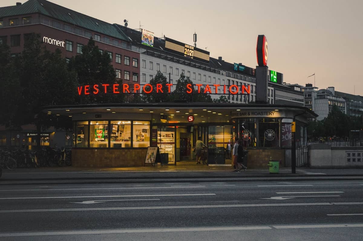 Kopenhag'da alacakaranlıkta Vesterport İstasyonu girişi, parlayan tabelalar ve arka planda şehir binaları ile.