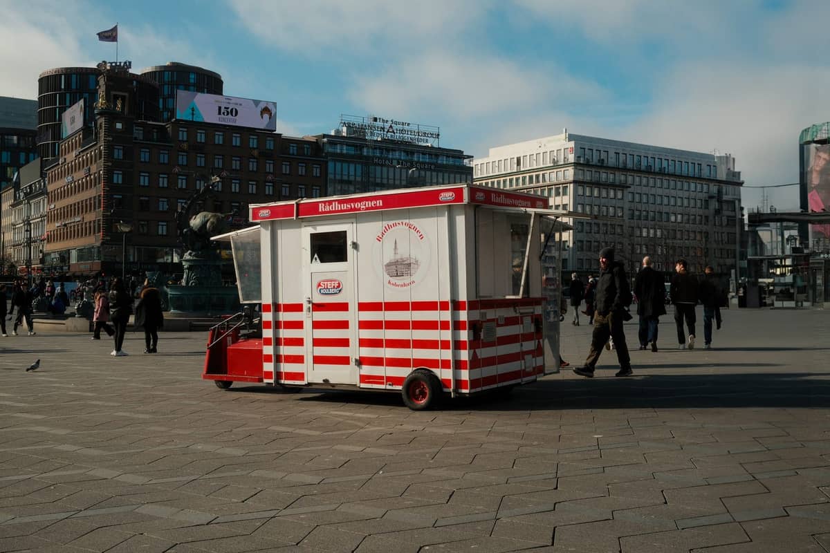 Clássico pølsevogn (carrinho de cachorro-quente) dinamarquês vermelho e branco em uma movimentada praça da cidade de Copenhague, com pedestres e edifícios urbanos ao fundo.