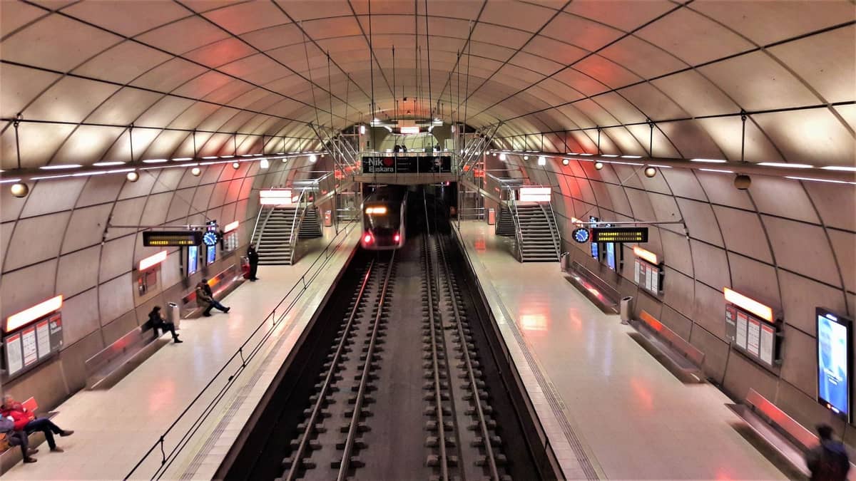 Estação de metrô subterrânea moderna em Copenhague com um trem chegando, pessoas esperando na plataforma e placas iluminadas.