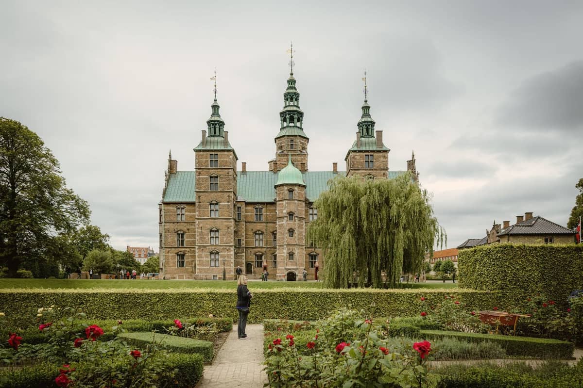 Vista do Castelo de Rosenborg visto dos seus jardins com uma pessoa caminhando por um caminho, sebes verdes e flores em primeiro plano.