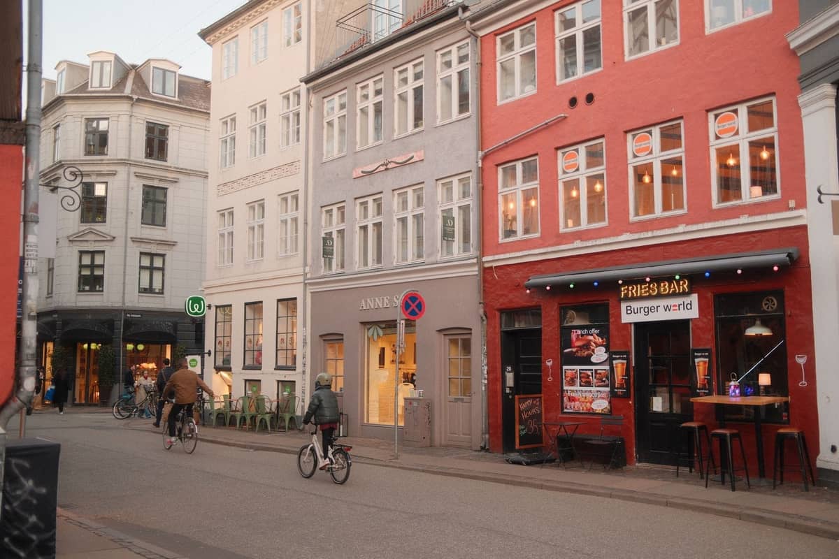Rua em Copenhague com edifícios coloridos, ciclistas e restaurantes baratos, sugerindo bairros locais acessíveis.