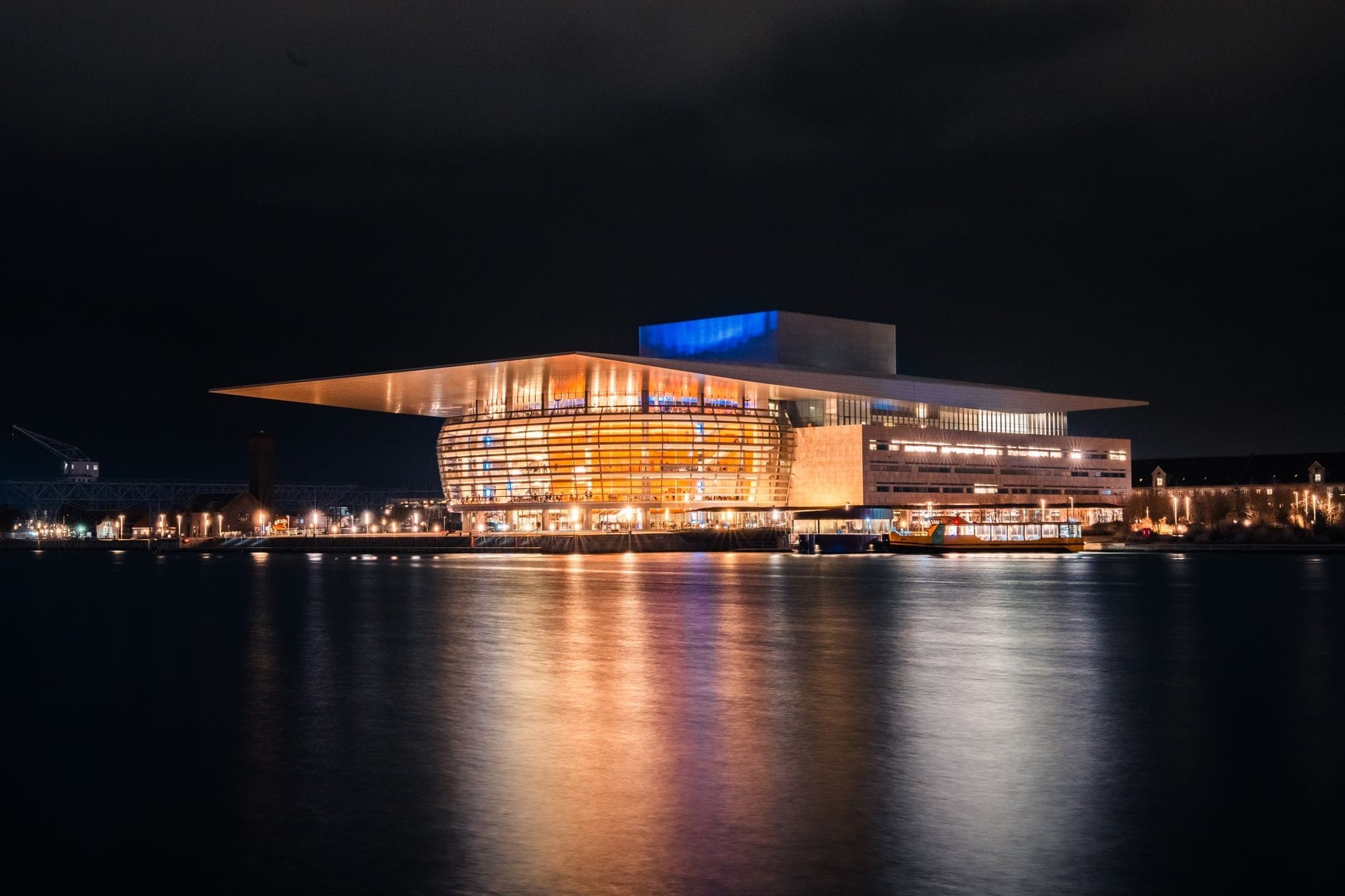 L'Opera di Copenaghen illuminata di notte, vista dal porto con i riflessi luminosi sull'acqua e una presenza architettonica moderna e suggestiva.