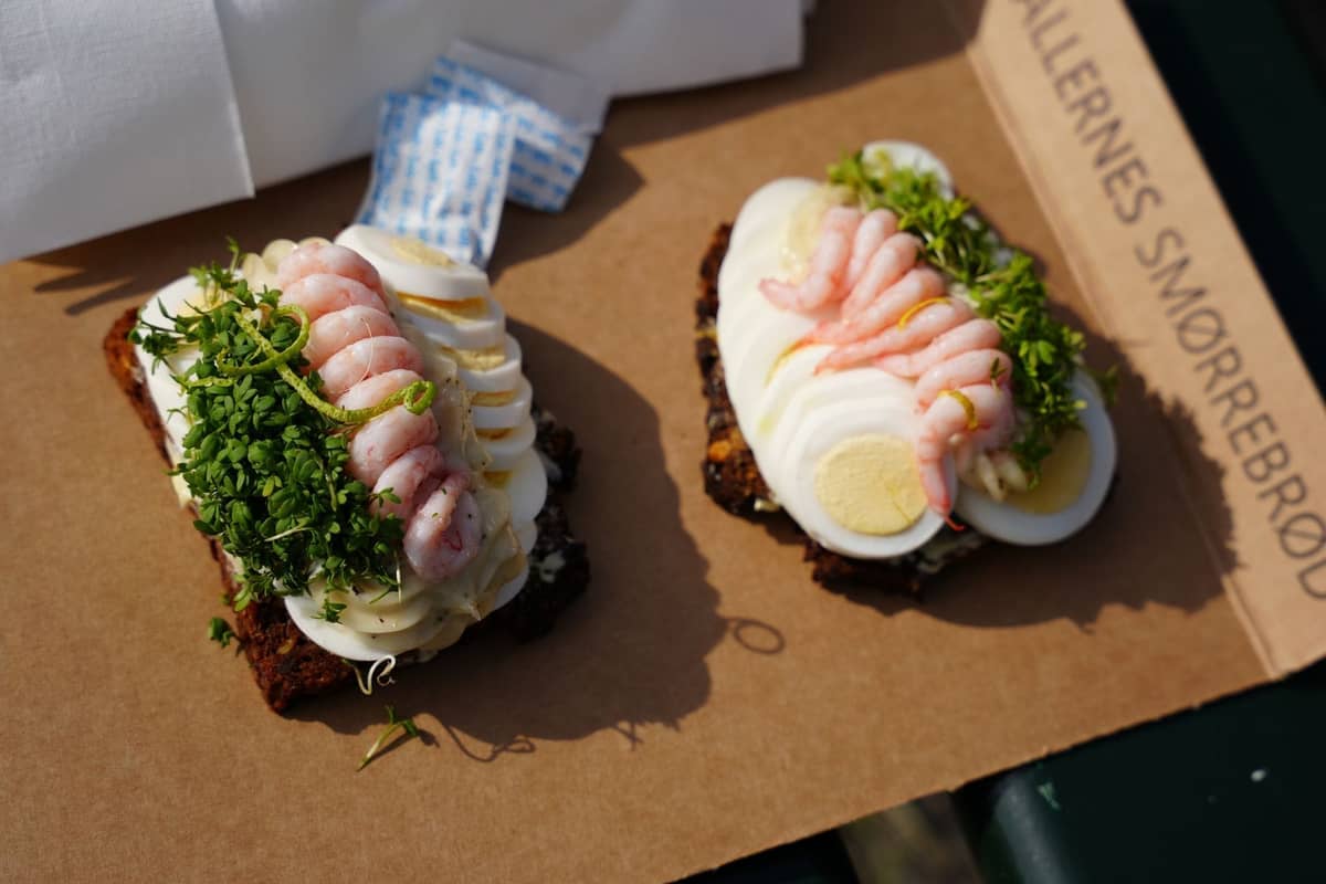 Dos piezas de smørrebrød clásico con gambas, huevo, mayonesa y hierbas sobre pan de centeno oscuro en una bandeja de cartón.