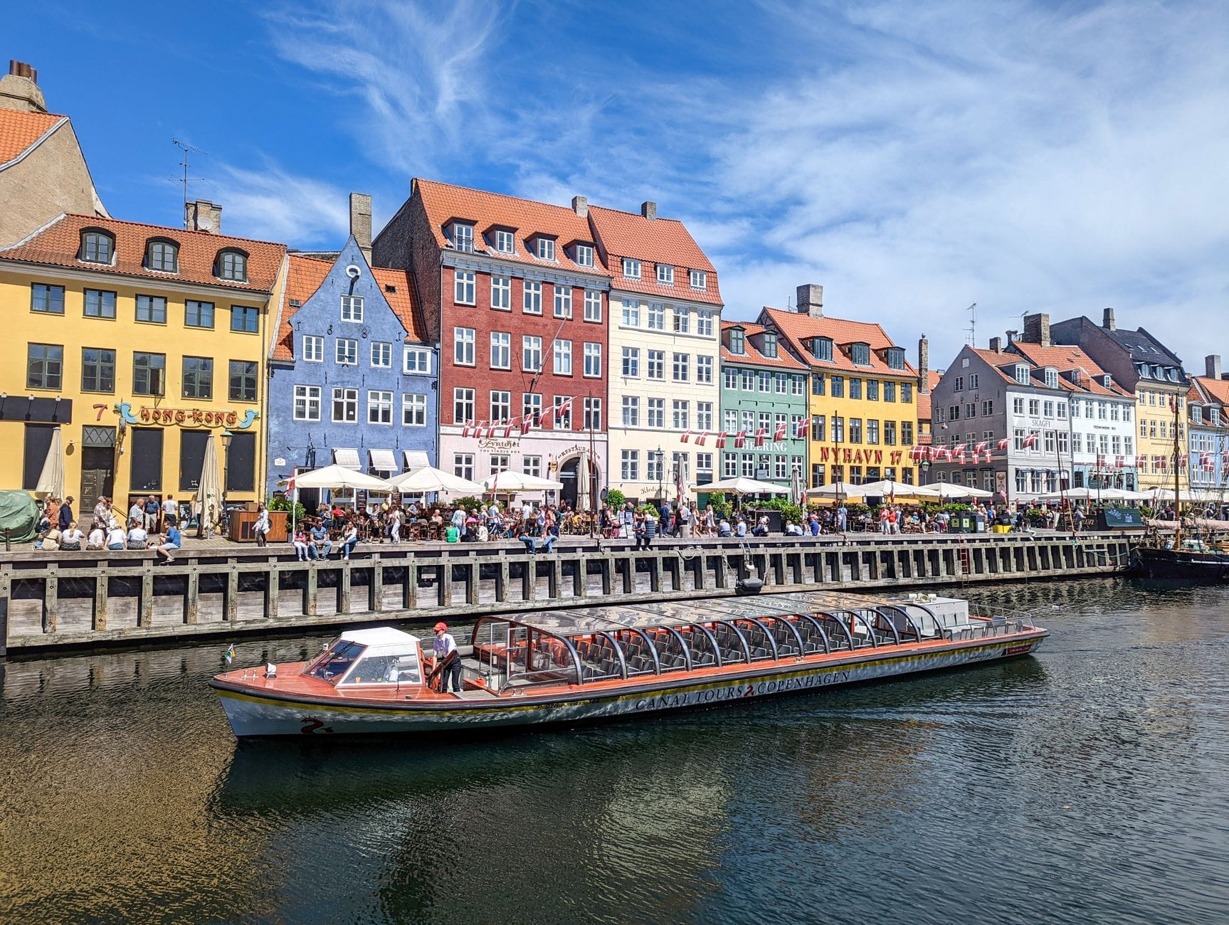 Casarões históricos e coloridos margeiam o canal de Nyhavn em Copenhague num dia ensolarado, com um barco turístico deslizando pelas águas e pessoas aproveitando os cafés ao ar livre.