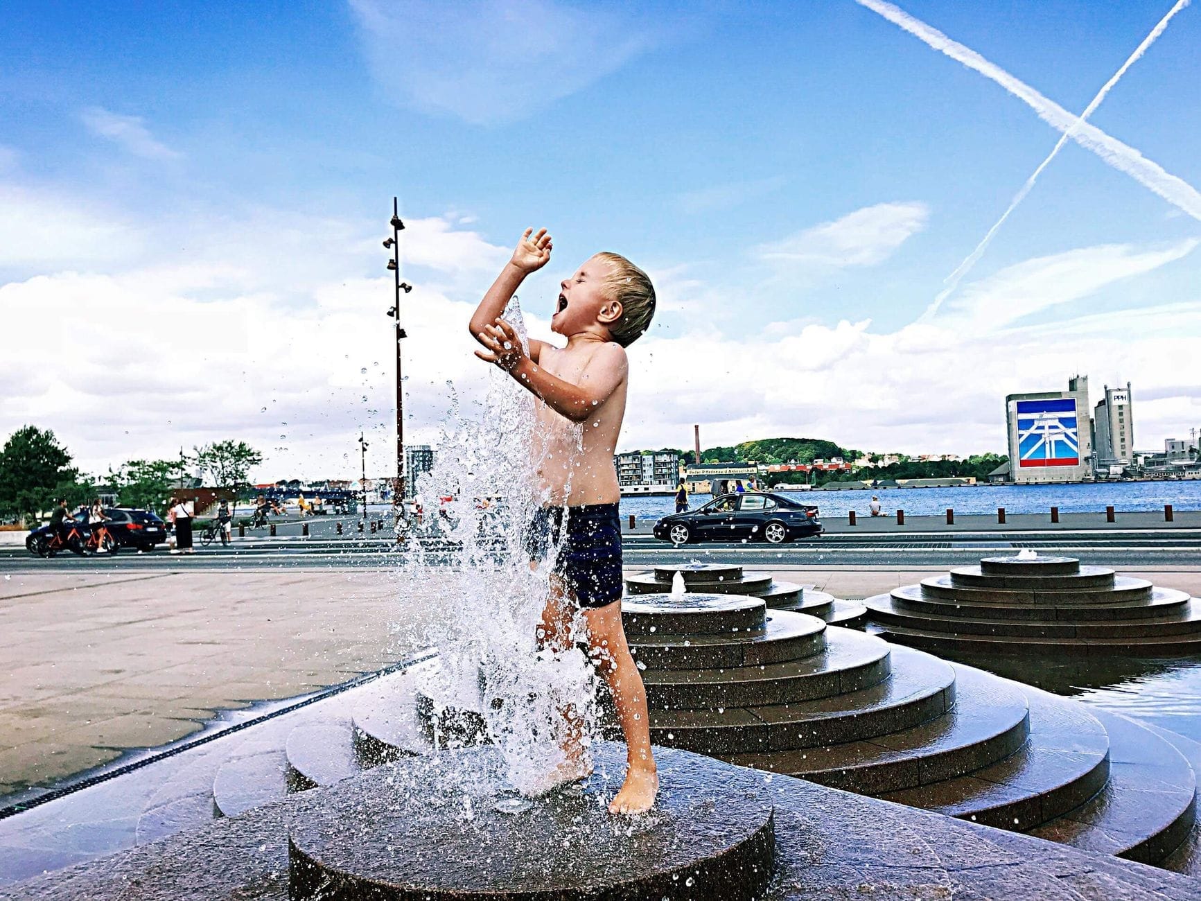 Uma criança pequena brincando animada em uma fonte urbana com água espirrando, em um dia ensolarado com um lindo cenário da cidade ao fundo.