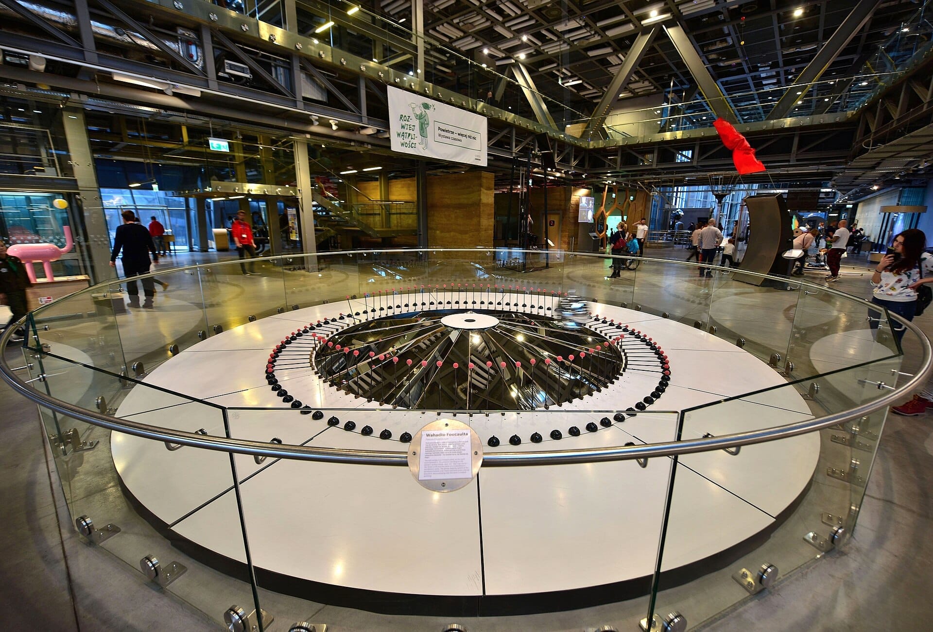 นิทรรศการลูกตุ้มฟูโกต์ขนาดใหญ่ล้อมรอบด้วยราวกระจกภายใน Copernicus Science Centre ที่ทันสมัยและสว่างไสว พร้อมนักท่องเที่ยวที่กำลังสำรวจพื้นที่