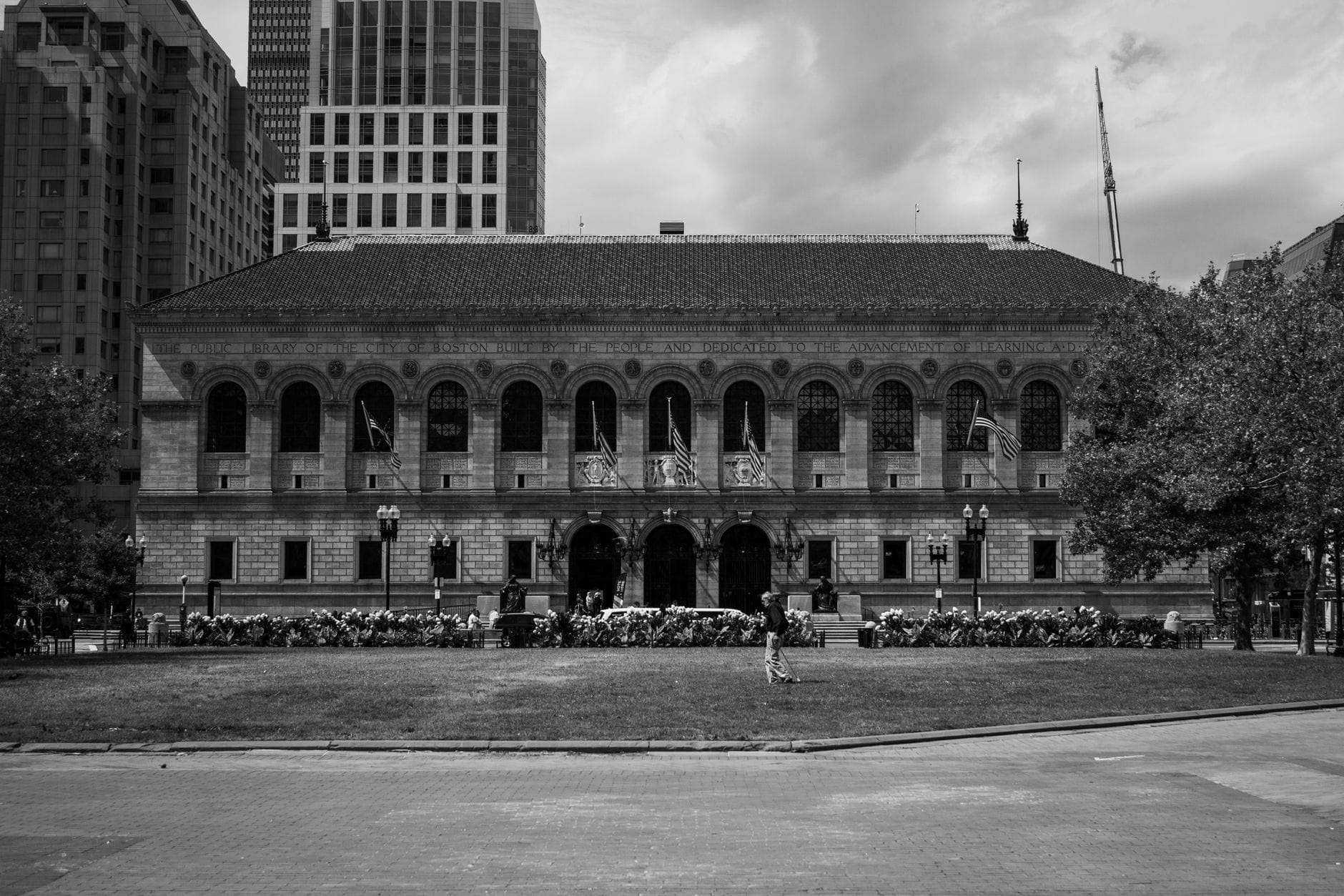 Vista frontale della storica Boston Public Library in Copley Square, circondata da grattacieli, con persone che camminano e verde in primo piano.