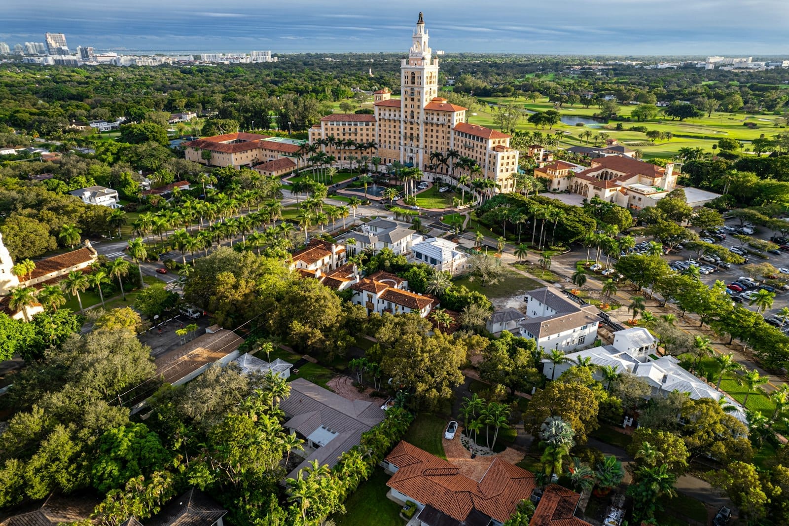ภาพถ่ายทางอากาศของ Coral Gables ที่มีโรงแรม Biltmore อันเก่าแก่ สนามกอล์ฟสีเขียว สถาปัตยกรรมเมดิเตอร์เรเนียน และบ้านเรียงรายใต้ร่มไม้ใหญ่ในวันที่แดดสดใส