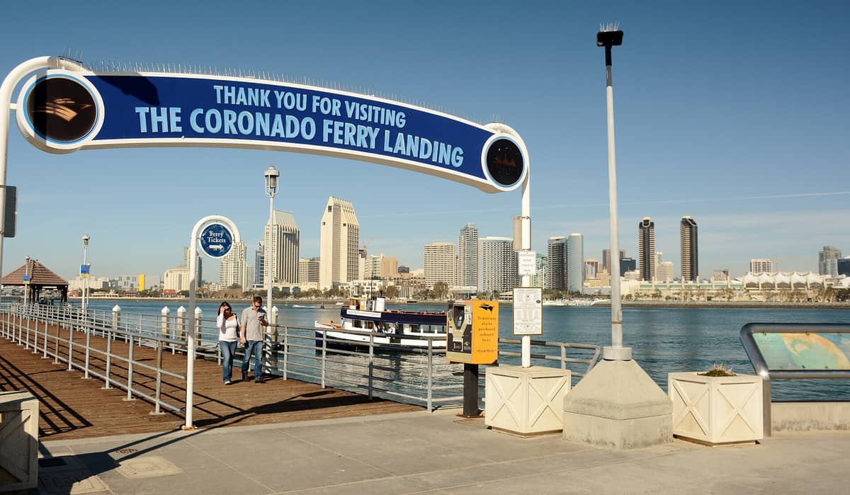 ป้ายทางเข้า Coronado Ferry Landing ทางเดินท่าเรือ จุดขึ้นเรือเฟอร์รี่ และวิวพาโนรามาเส้นขอบฟ้าแซนดีเอโก อ่าว และท้องฟ้าใส ๆ