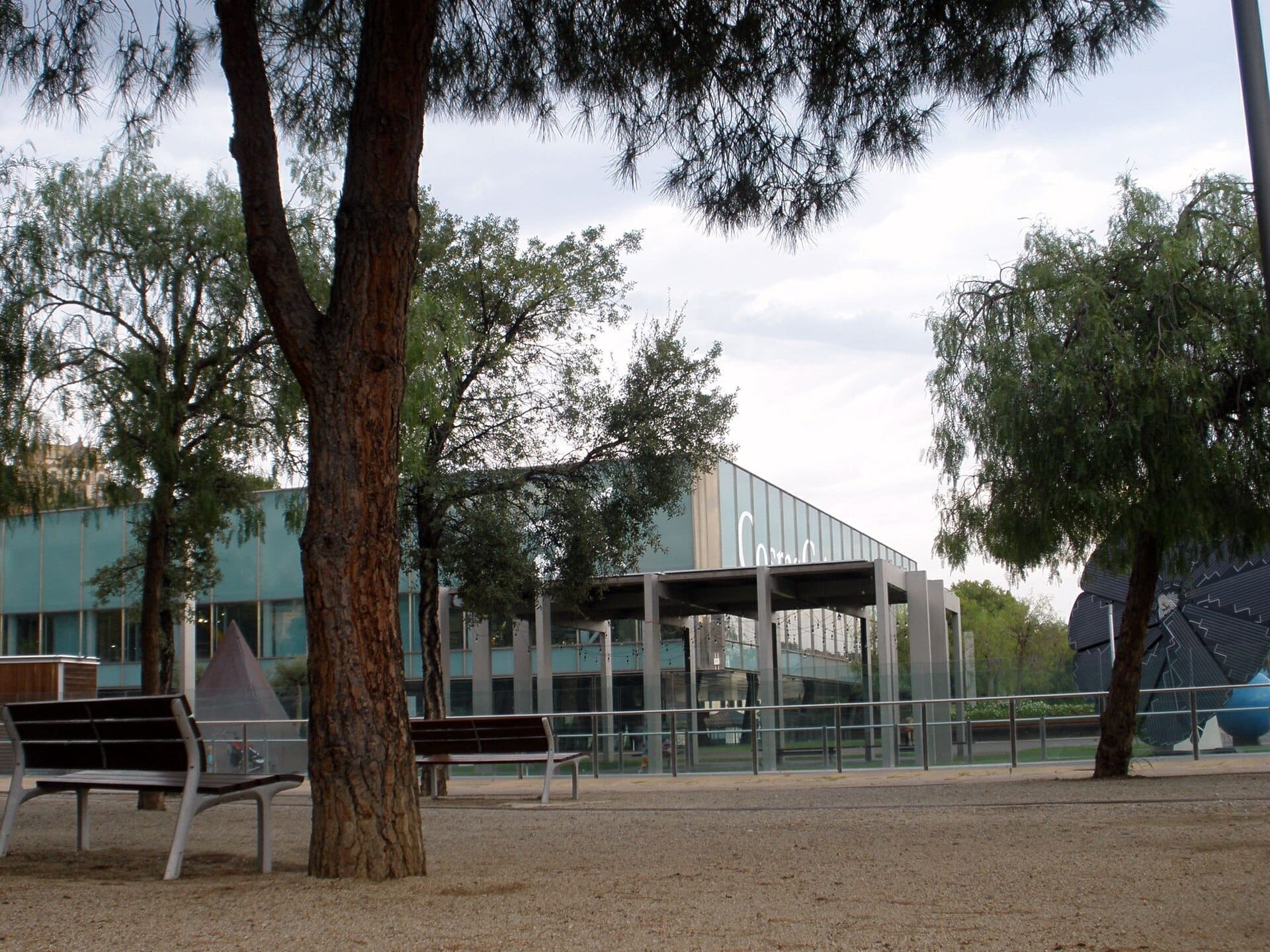 CosmoCaixa Barcelona exterior view with modern glass facade, surrounded by trees and benches on a cloudy day.