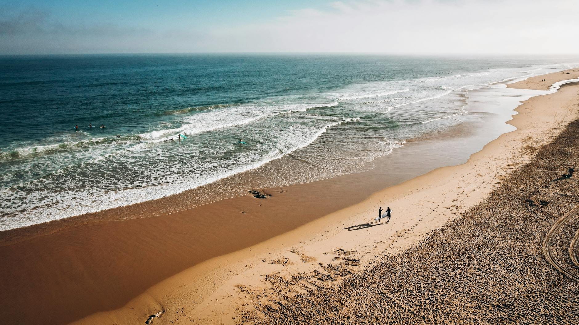 Vista aerea di una lunga spiaggia sabbiosa con onde turchesi e alcune persone che camminano lungo la riva sotto un cielo luminoso.