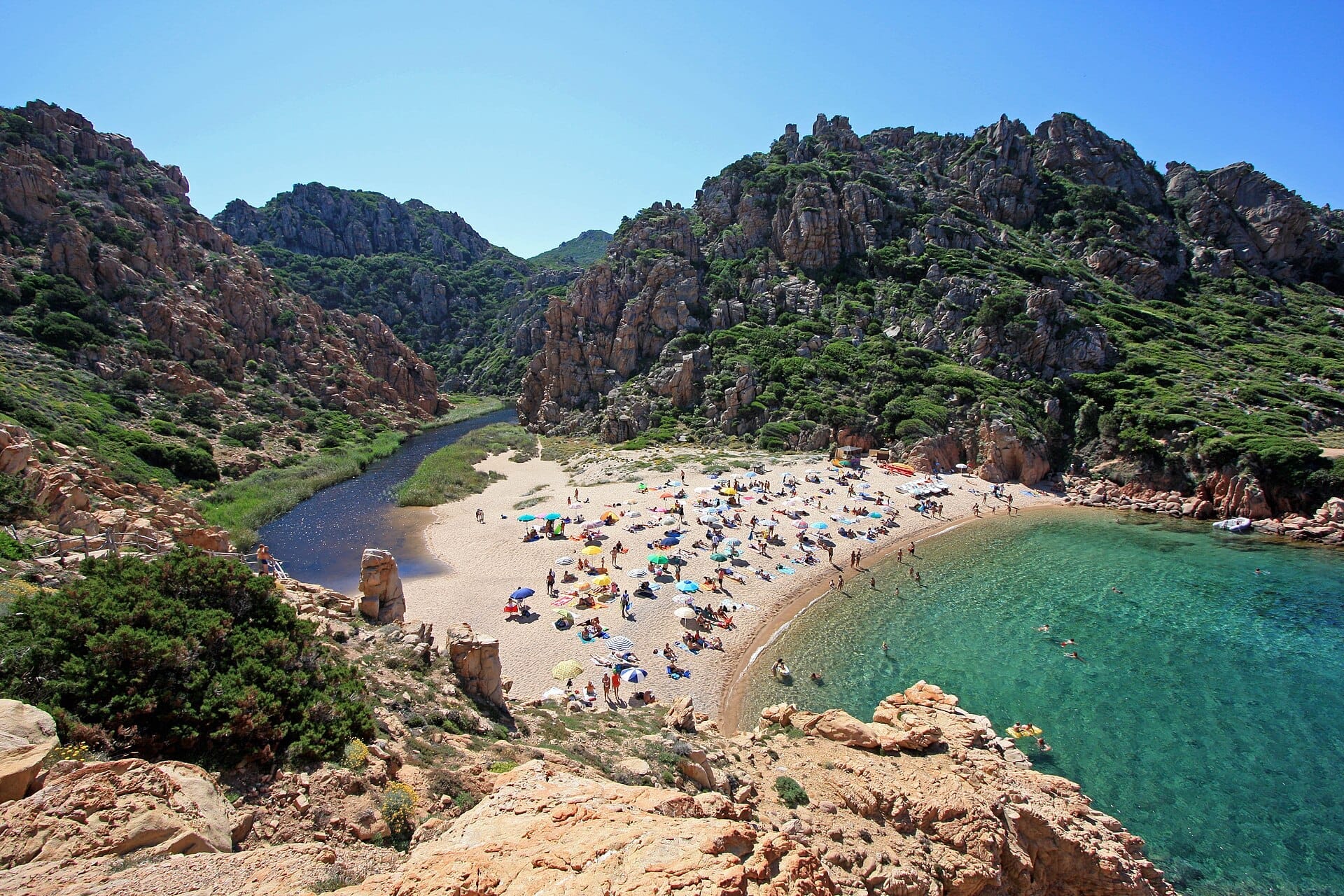 Sandstrand an der Costa Paradiso, umgeben von rauen roten Granitfelsen, klarem türkisfarbenem Wasser und Sonnenbadenden mit bunten Sonnenschirmen an einem strahlend sonnigen Tag.
