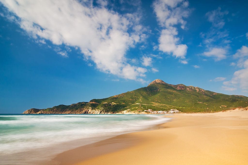 Breiter Sandstrand und türkisfarbene Wellen der Costa Verde auf Sardinien, im Hintergrund grüne Hügel und einige kleine weiße Gebäude.