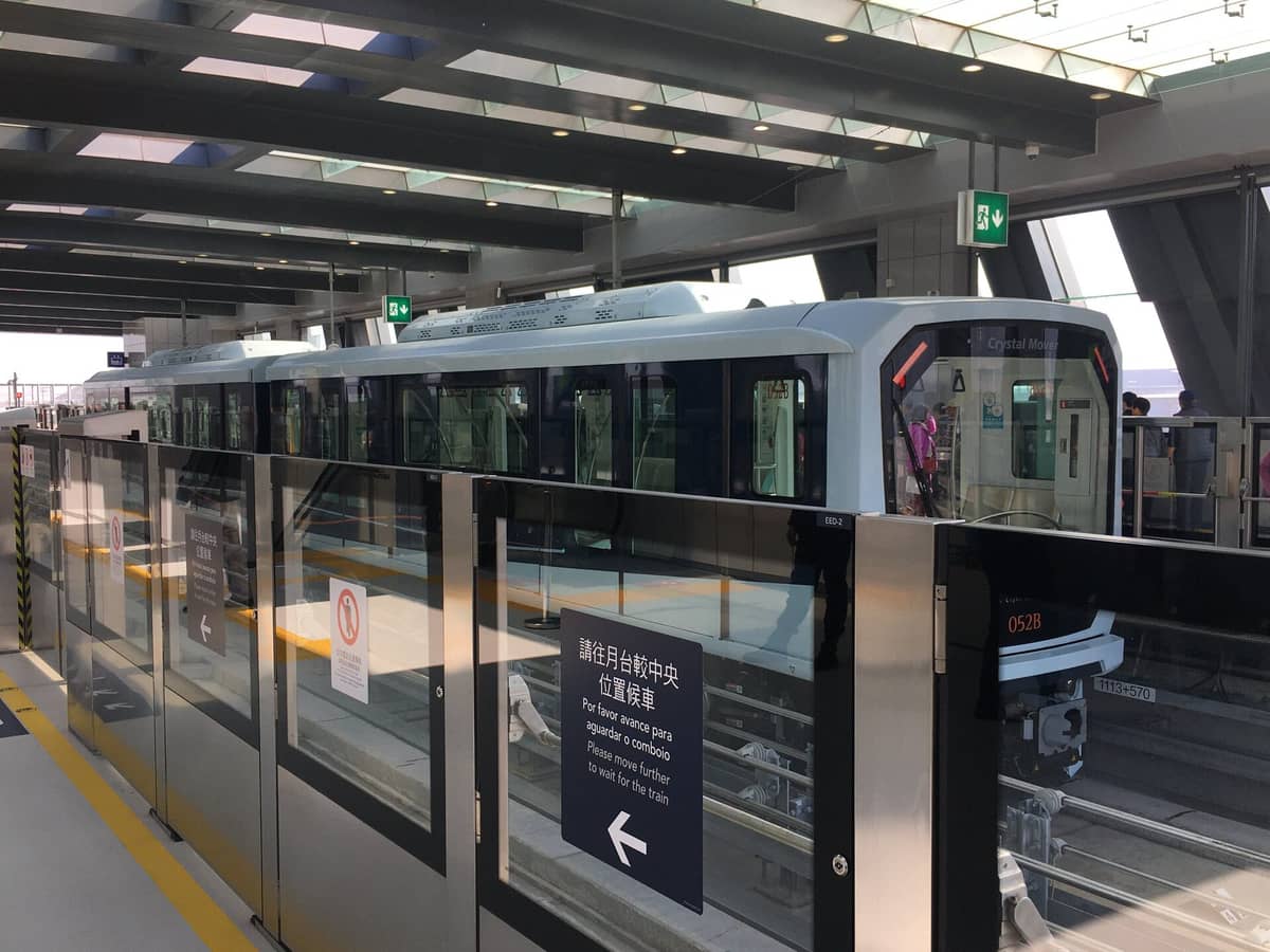 Ein moderner, erhöhter LRT-Bahnhof in Macau mit einem Crystal Mover-Zug, der am Bahnsteig hält; Glastüren und Deckenbeleuchtung sind sichtbar.