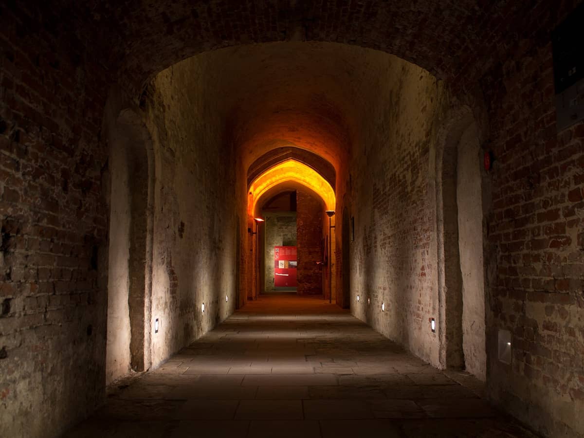 Longo corredor de tijolos no interior do Sítio Arqueológico do Palácio de Coudenberg, iluminado com luz quente e conduzindo a uma porta arqueada em Bruxelas.