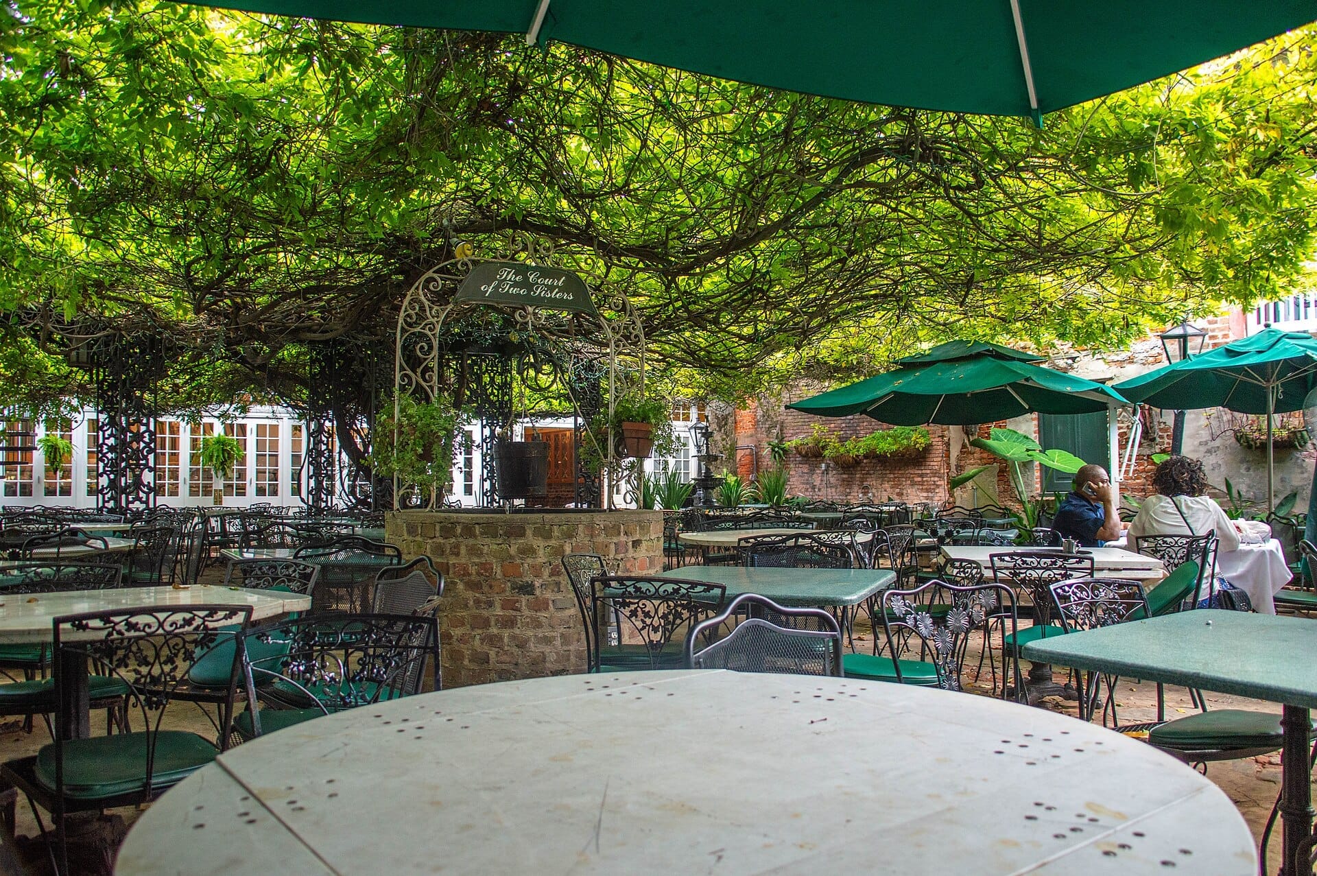 Area pranzo nel cortile del The Court of Two Sisters, con tavoli in ferro battuto, ombrelloni verdi, pozzo in mattoni e rigogliosa vegetazione in alto.