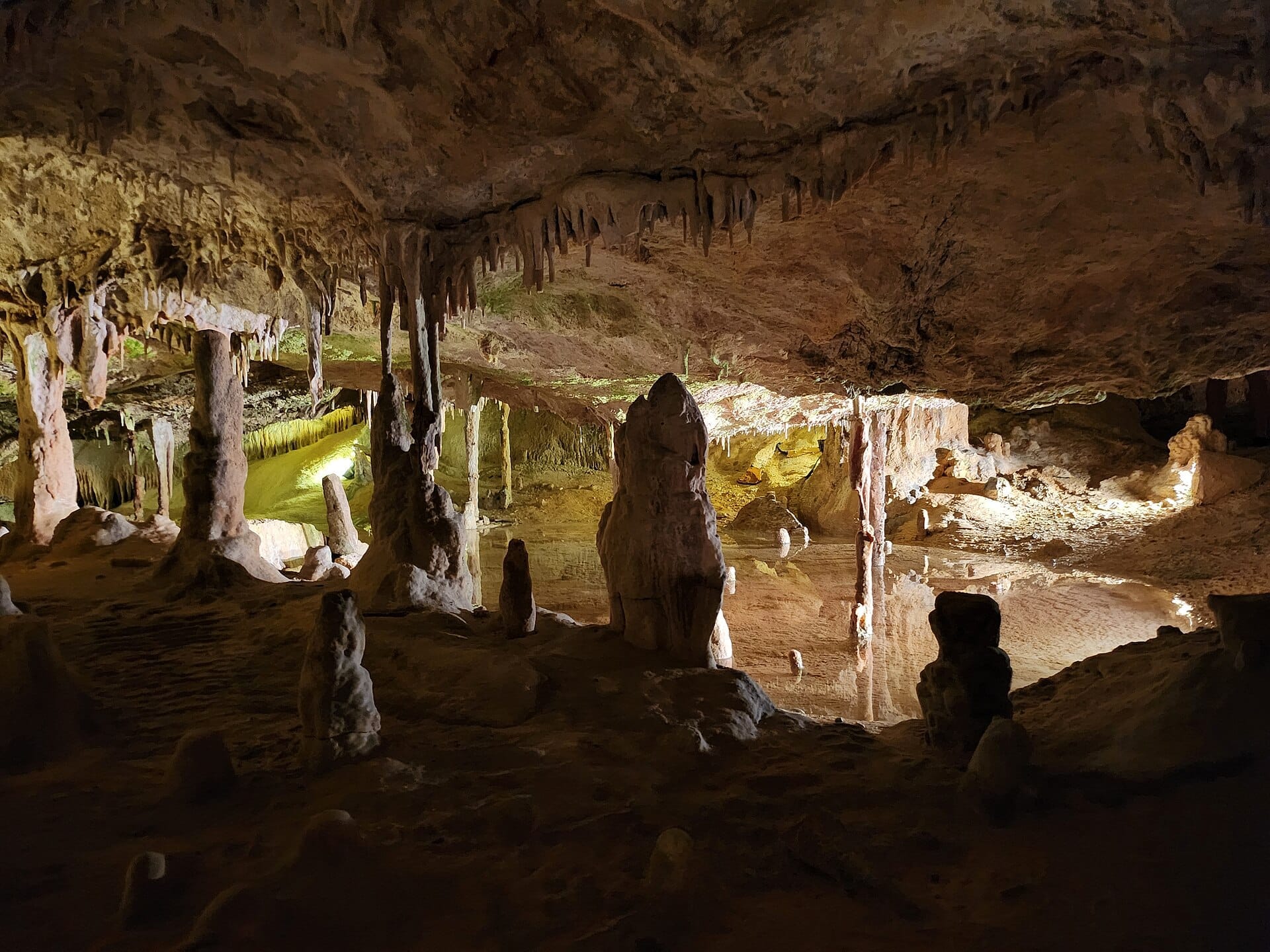 Vista interna della Cova de Can Marçà con stalattiti rocciose, alte colonne di grotta e una piscina sotterranea riflettente illuminata da luci calde.