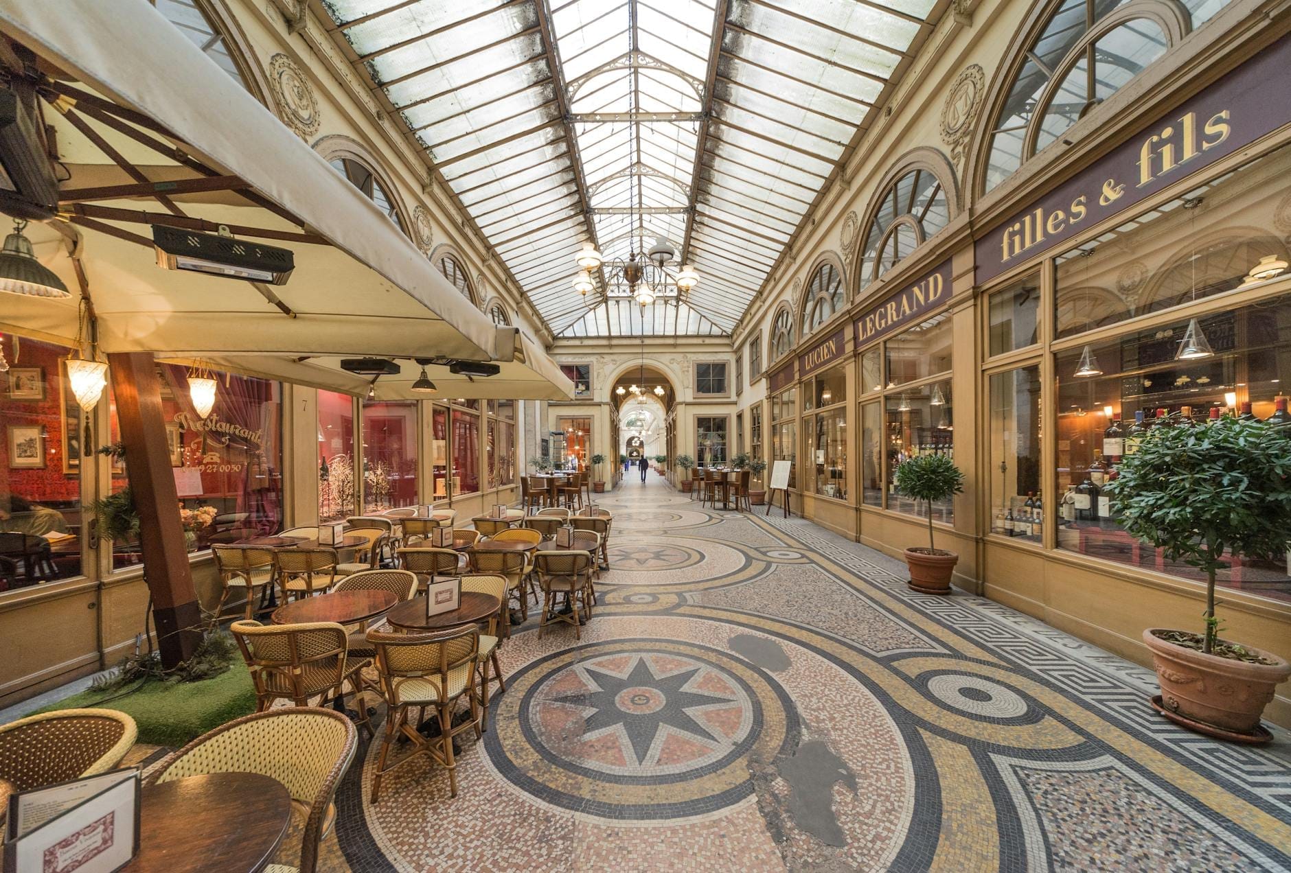 Vista interna di un passage coperto parigino con pavimenti a mosaico, tetto in vetro, eleganti vetrine e tavolini di un caffè lungo la galleria.
