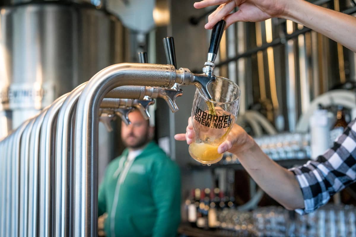Ein Barkeeper zapft ein Pintglas an glänzenden Bierhähnen in einem modernen Brauerei-Taproom, im Hintergrund sind Gärtanks und Gläser zu sehen.