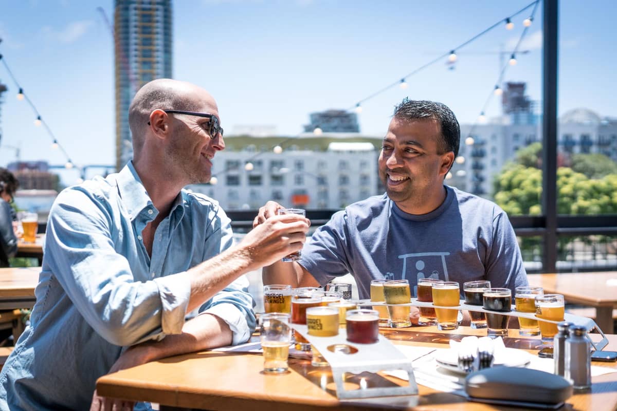 Zwei Personen stoßen mit Biergläsern an einem sonnenbeschienenen Tisch auf einer Außenterrasse an, im Hintergrund sind Beer-Flight-Boards, Stadtgebäude und Lichterketten zu sehen.